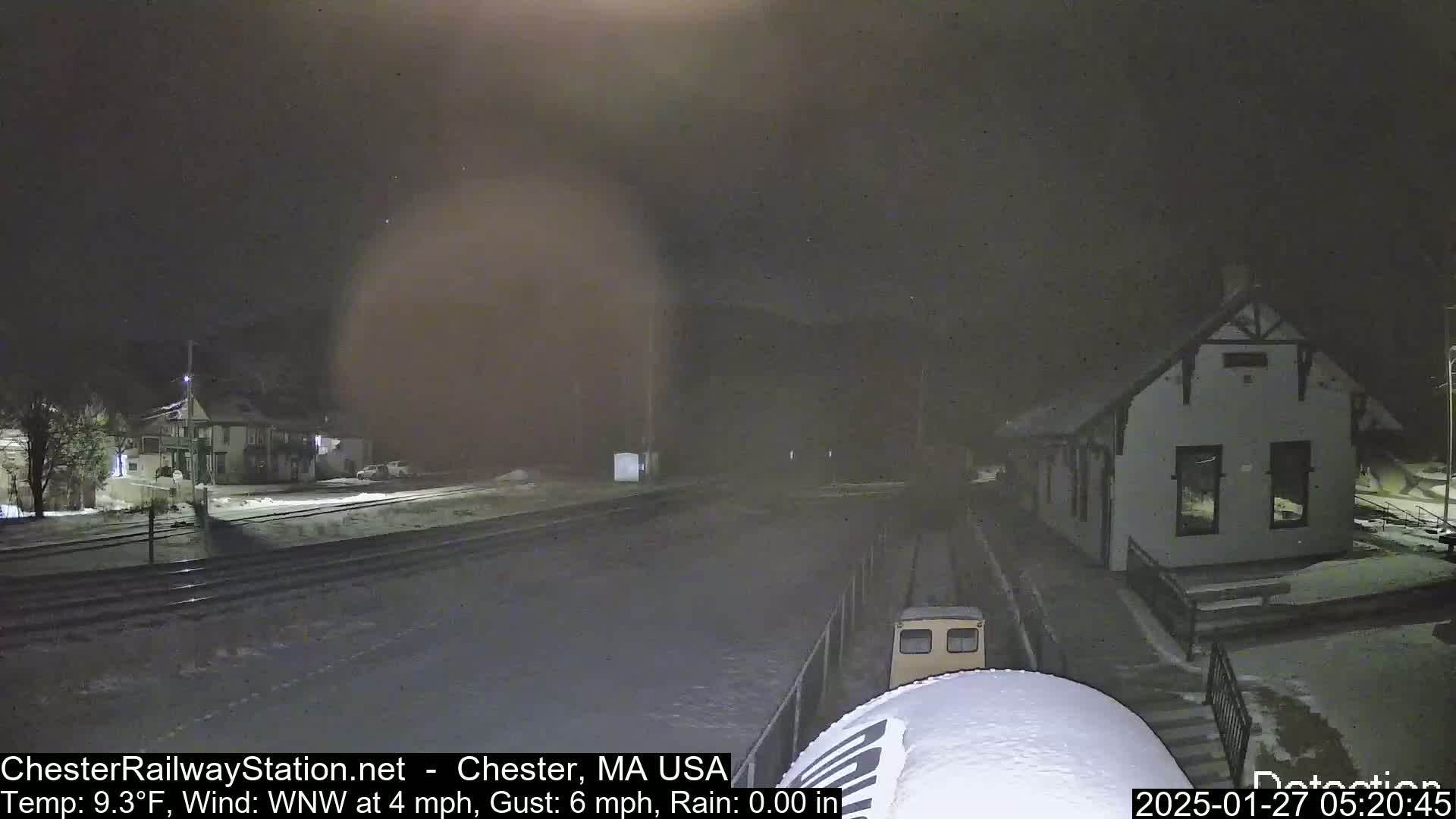 A nighttime view of a snow-covered train station and tracks with a small vehicle parked nearby, under a dark, slightly hazy sky.