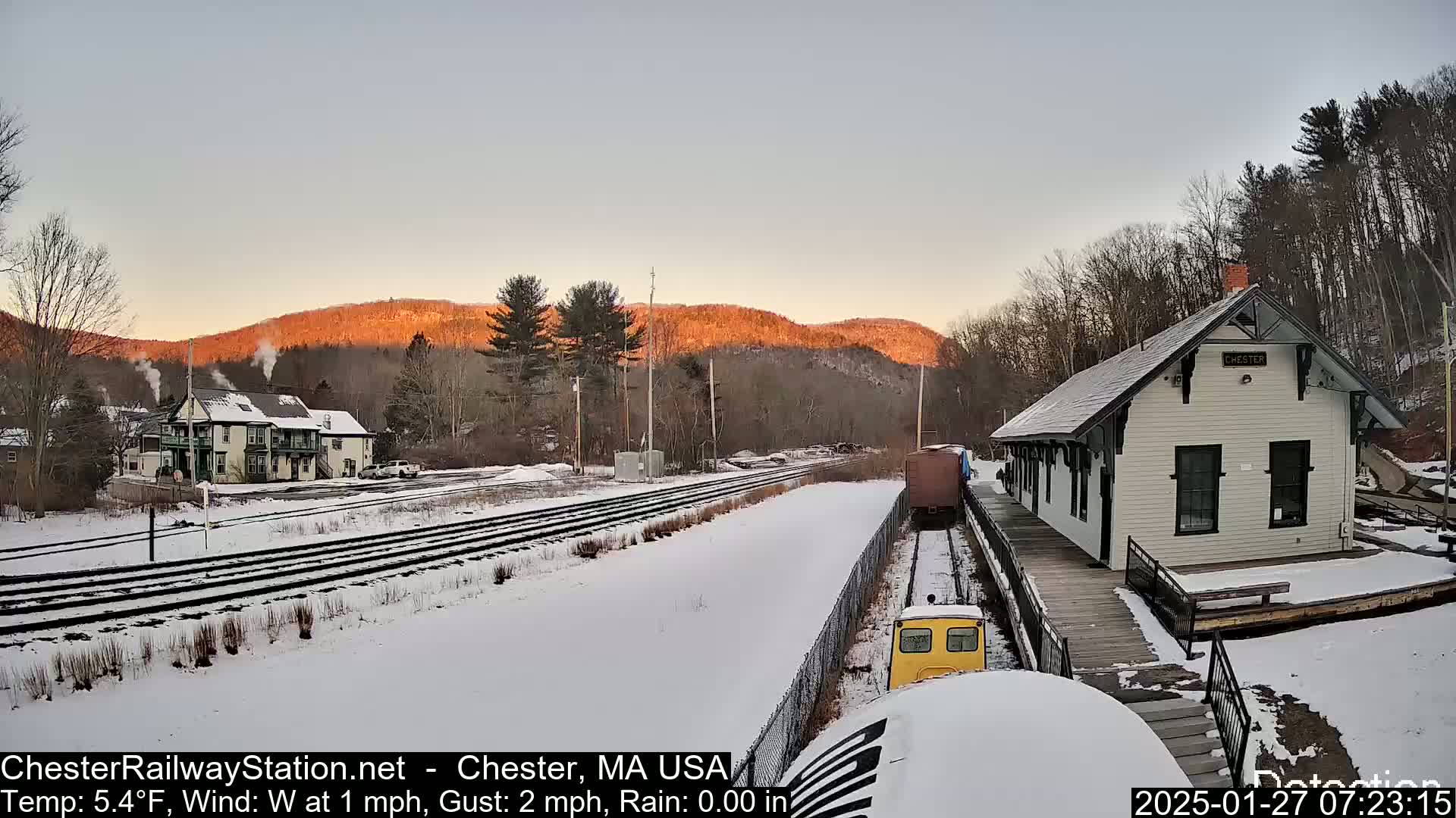 A snow-covered train station and rail yard are situated in front of a sunlit mountain range at sunrise.