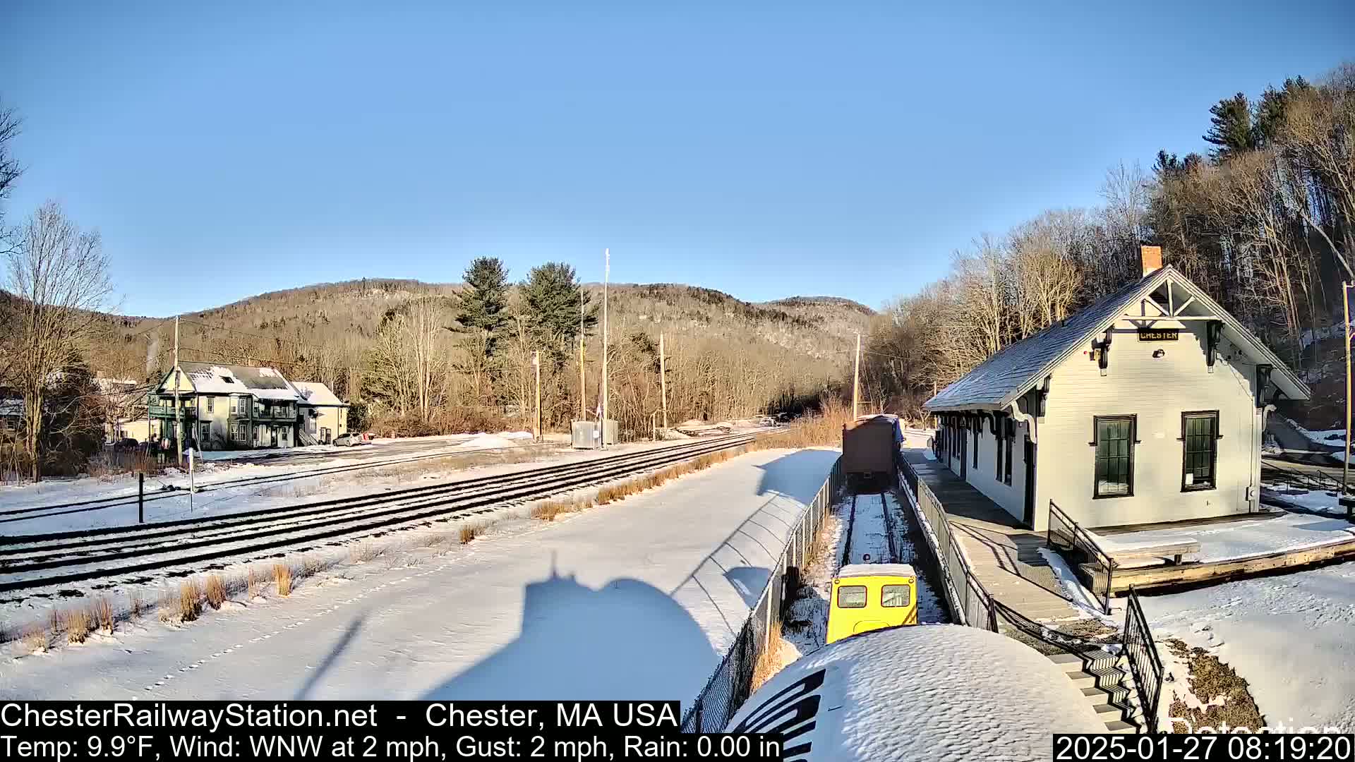 A snow-covered train station and railway track are situated in a valley, with a small yellow train car visible in the foreground under a clear, sunny sky.