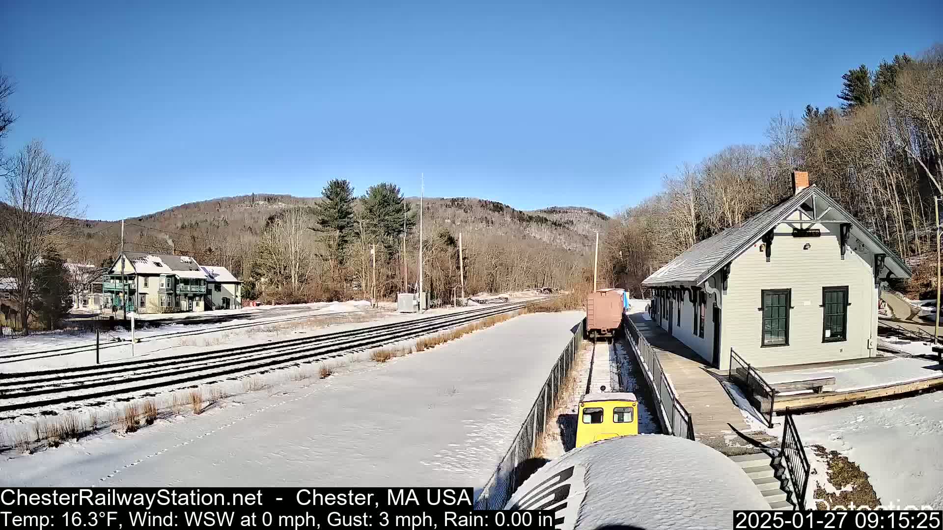 A snow-covered train station and tracks are situated in a valley under a clear, sunny sky.