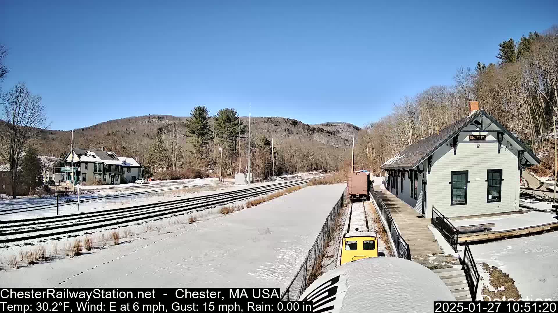 A small yellow train approaches a snow-covered train station on a sunny day with a light east wind.