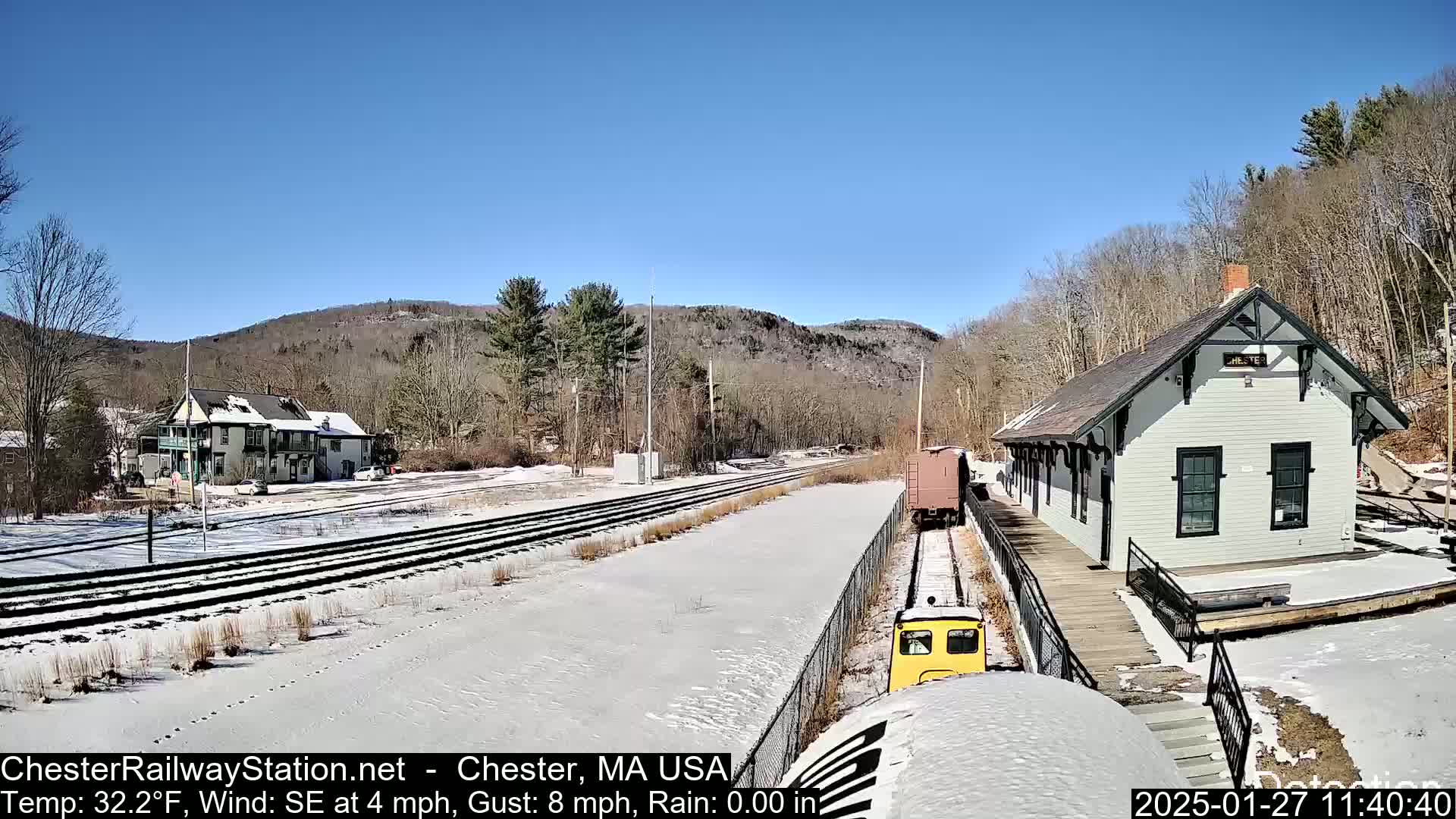 A small yellow train approaches a snow-covered train station on a sunny day with a light southeast wind.