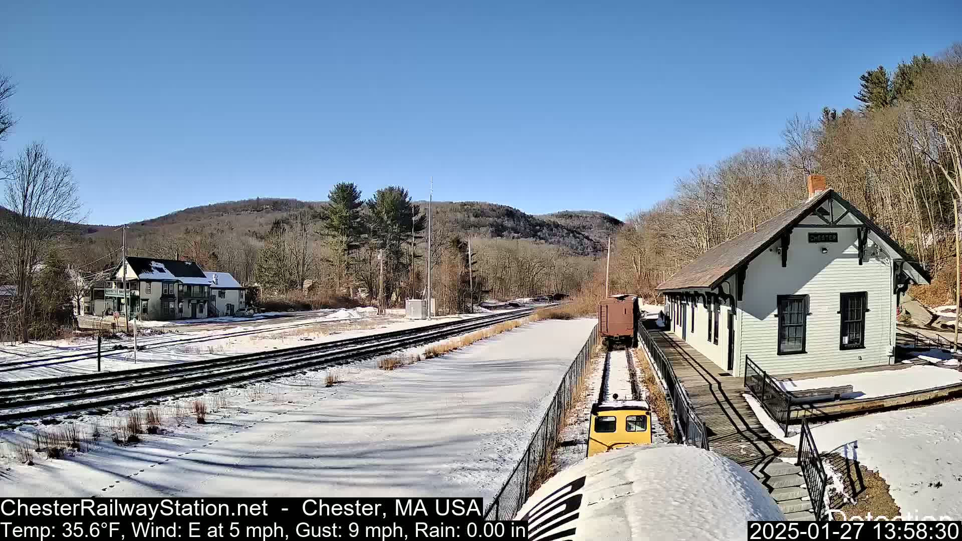 A small yellow train approaches a snow-covered train station on a sunny day with a light east wind.