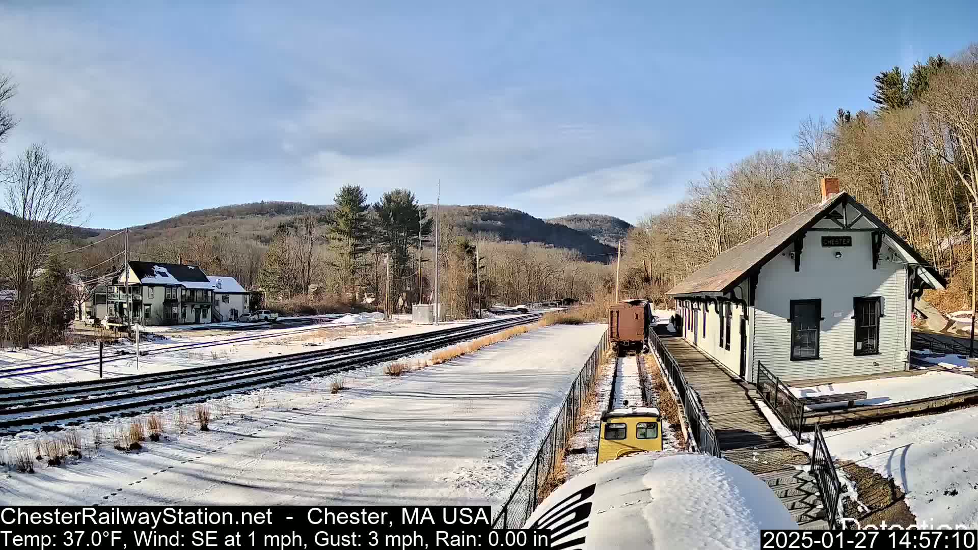 A snow-covered train station and tracks sit beside a small building, with hills and woods visible in the background under a mostly clear sky.