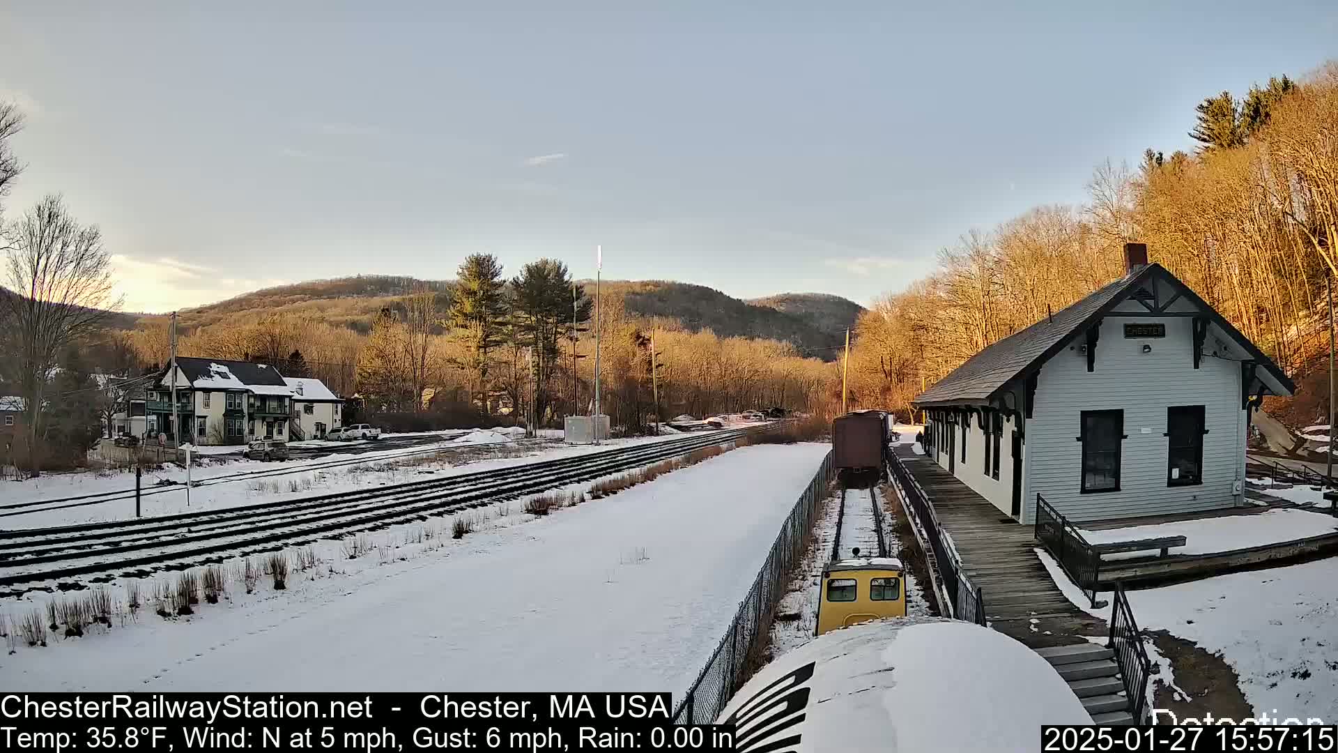 A snow-covered railway station with a small train approaching, nestled among bare trees and hills under a partly sunny sky.