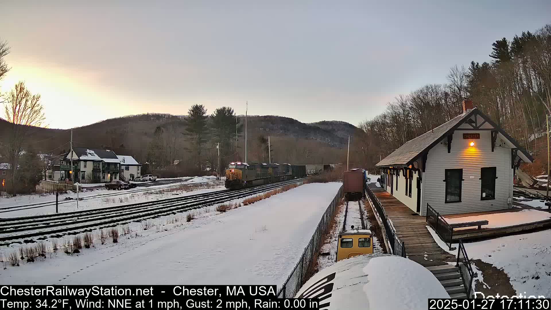 A freight train passes a snow-covered train station at sunset under a clear sky.