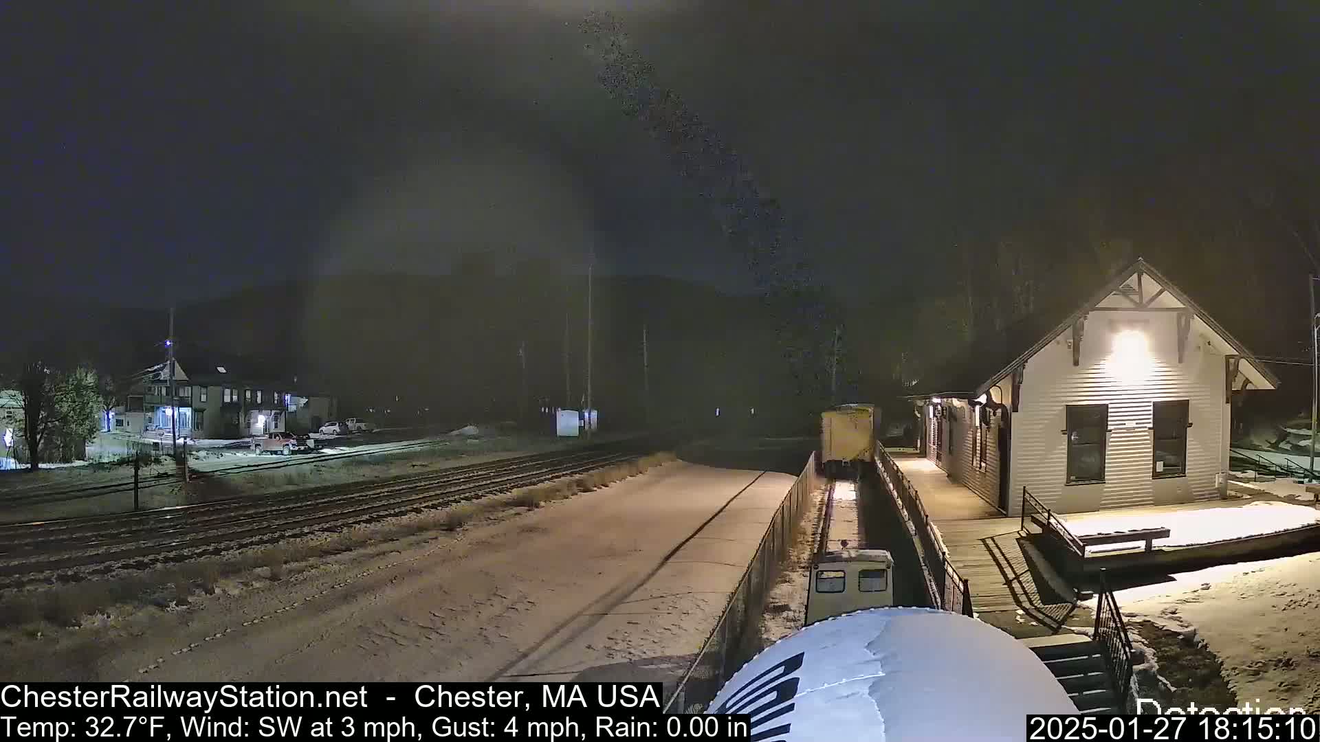 A nighttime scene of a snow-covered train platform next to a small building with a train car visible nearby, under a dark sky with a southwest wind.