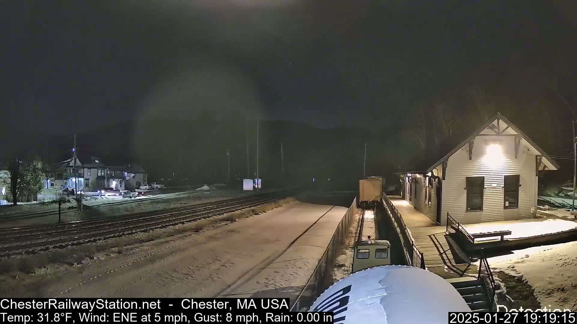 A nighttime snow-covered train station scene shows a train car on the tracks next to a building, with cars parked across the street.