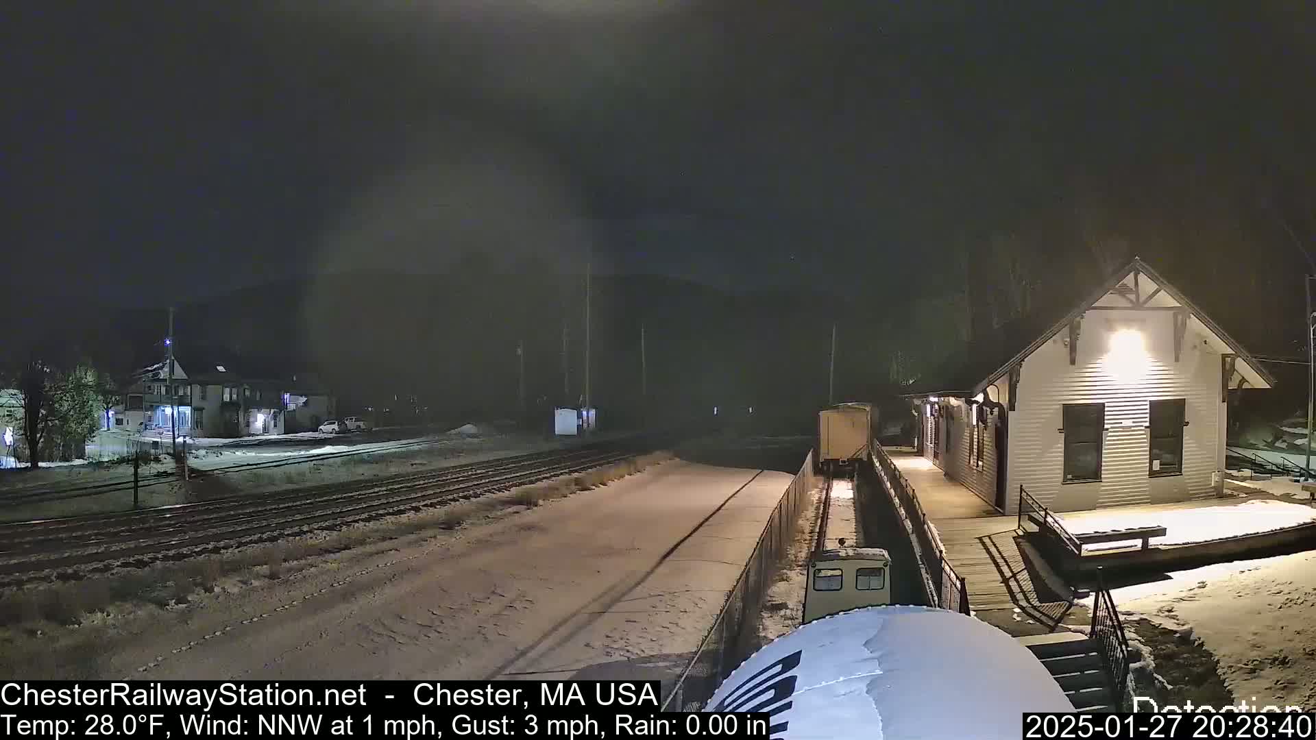 A snow-covered train station at night with a train car partially visible on the tracks in front of it, under a dark sky with light snow falling.