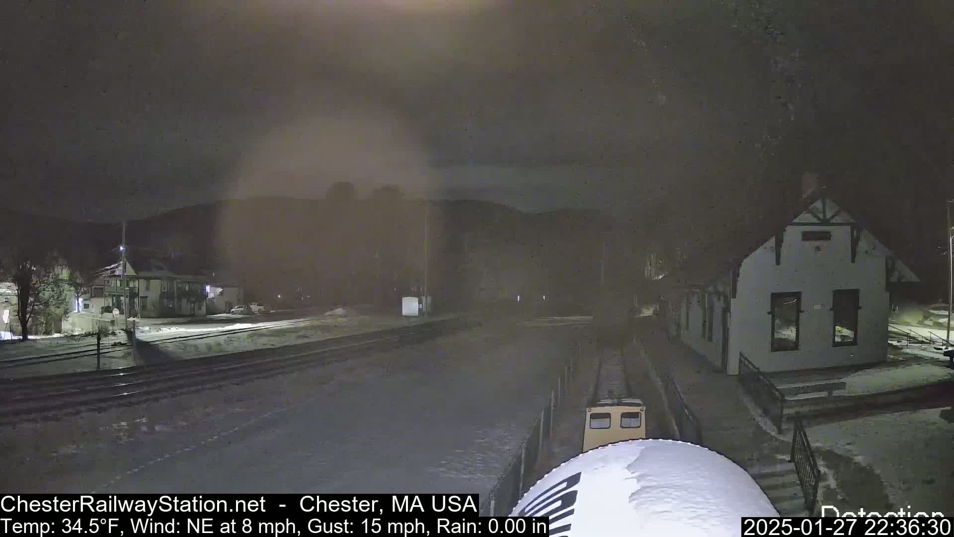 A nighttime view of a snow-covered train station and tracks, with a small train car partially visible, under a dark, cloudy sky with a northeast wind.