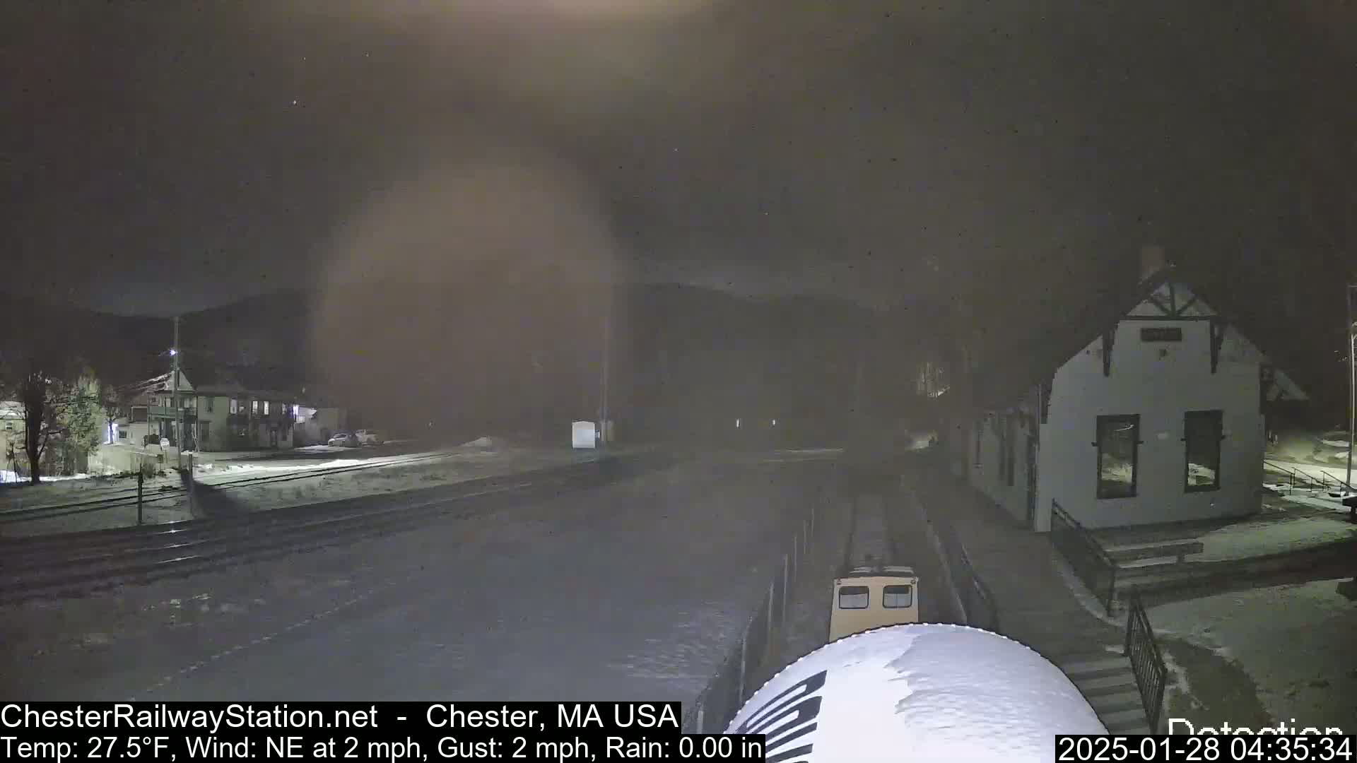 A small train is visible on a snow-covered railway track at night near a building, under a dark sky with light rain or snow.