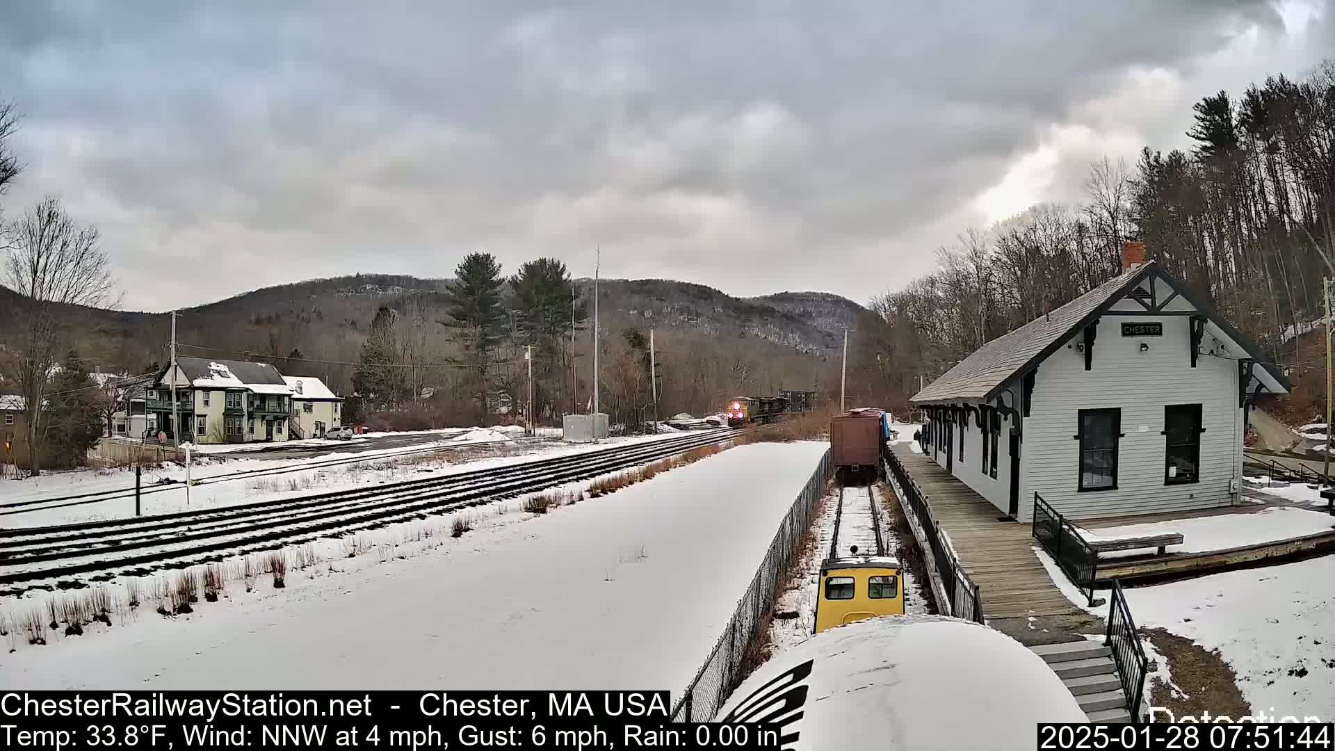 A train passes a snow-covered train station in a hilly landscape under a cloudy sky.