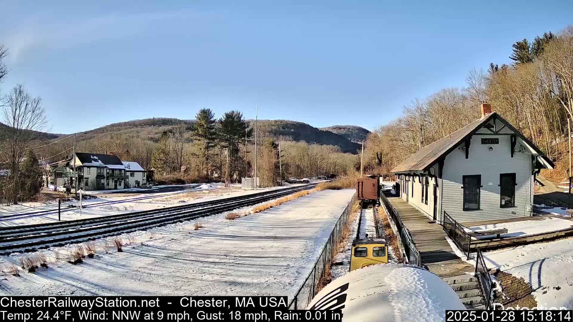 A train approaches a snow-covered railway station and tracks, with a building and hills visible in the background under a clear, sunny sky.