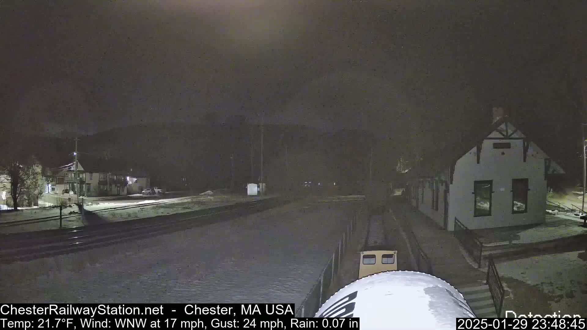 A nighttime scene of a train partially visible in the foreground at a snow-covered train station under rainy conditions with a wind of 17 mph gusting to 24 mph, and a temperature of 21.7°F.
