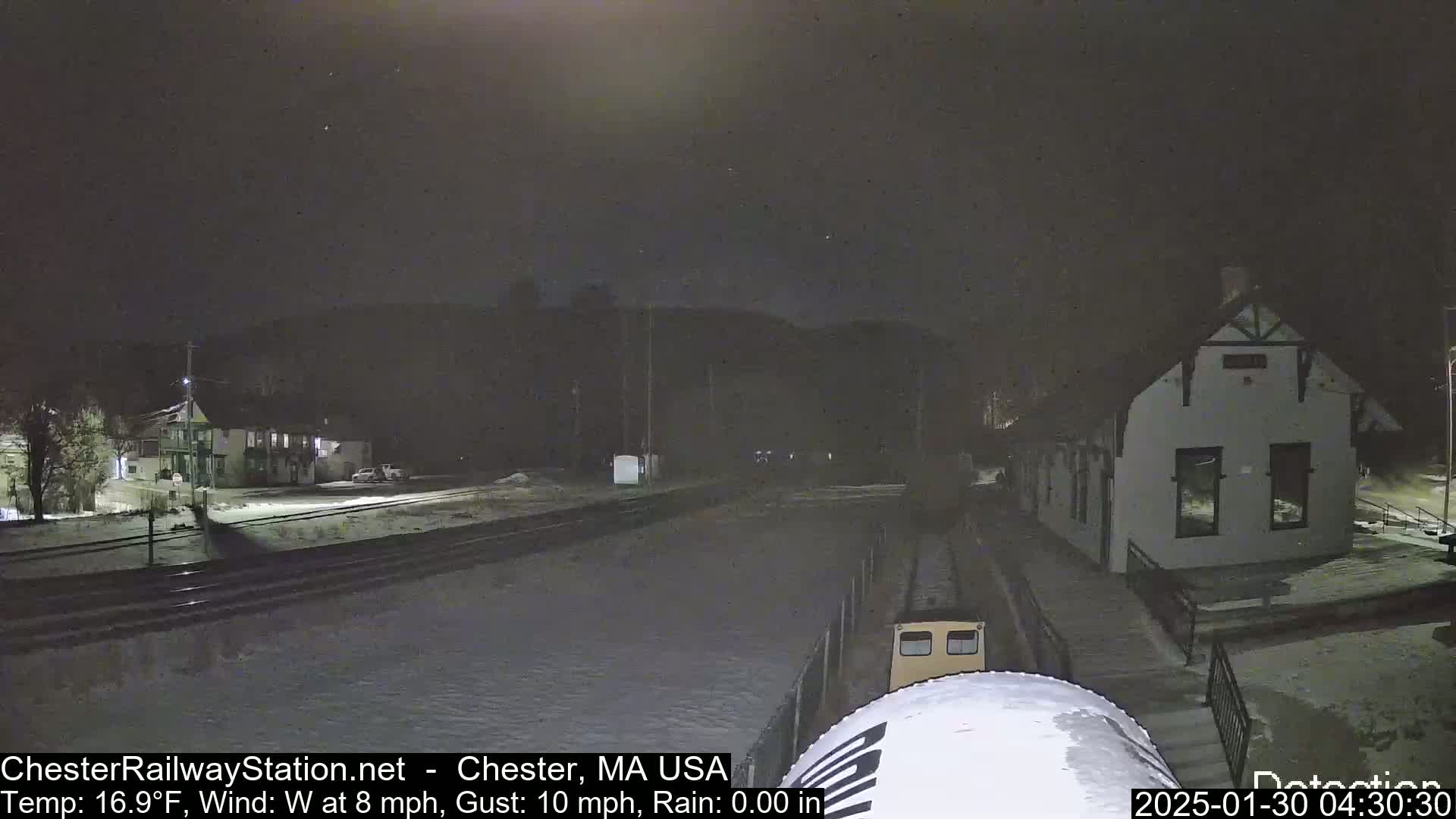 A nighttime view of a train approaching a snow-covered train station under a dark sky with a west wind.