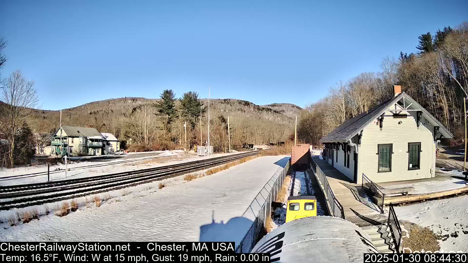 A snow-covered train station and tracks sit against a backdrop of a wooded hillside under a clear blue sky.