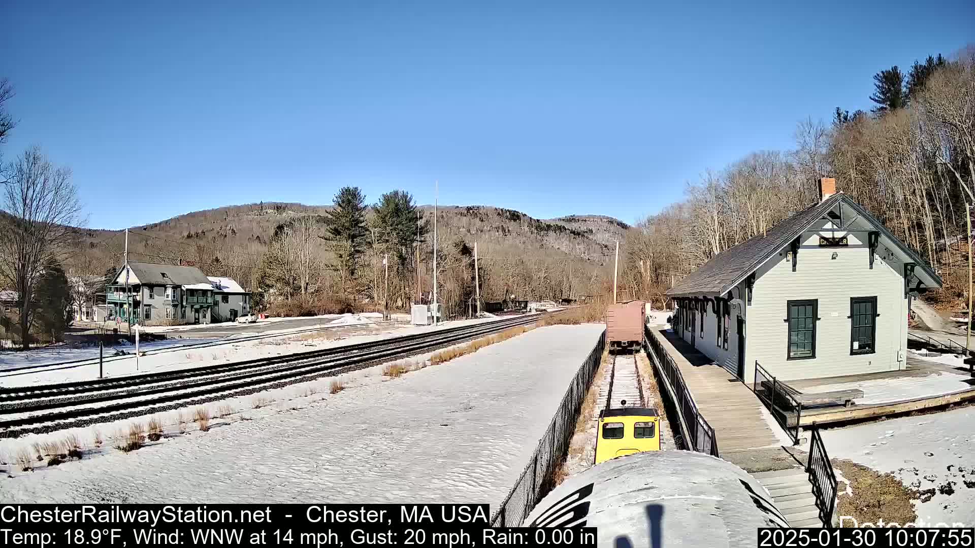 A small yellow train approaches a snow-covered train station in a sunny, cold, windy setting, with bare trees and hills in the background.