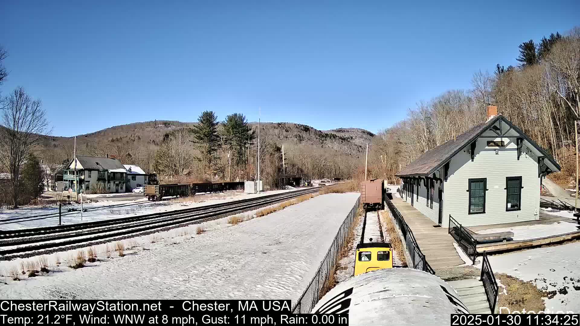 A small yellow train approaches a snow-covered train station, nestled beside a building and a hillside under a clear blue sky.