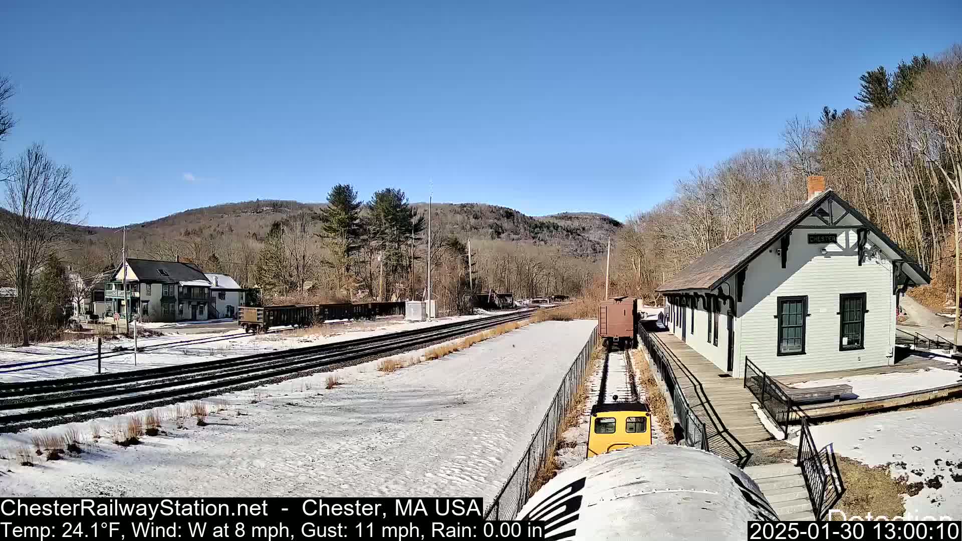 A small yellow train approaches a white train station on a snow-covered railway line under a clear blue sky, with hills and trees in the background.