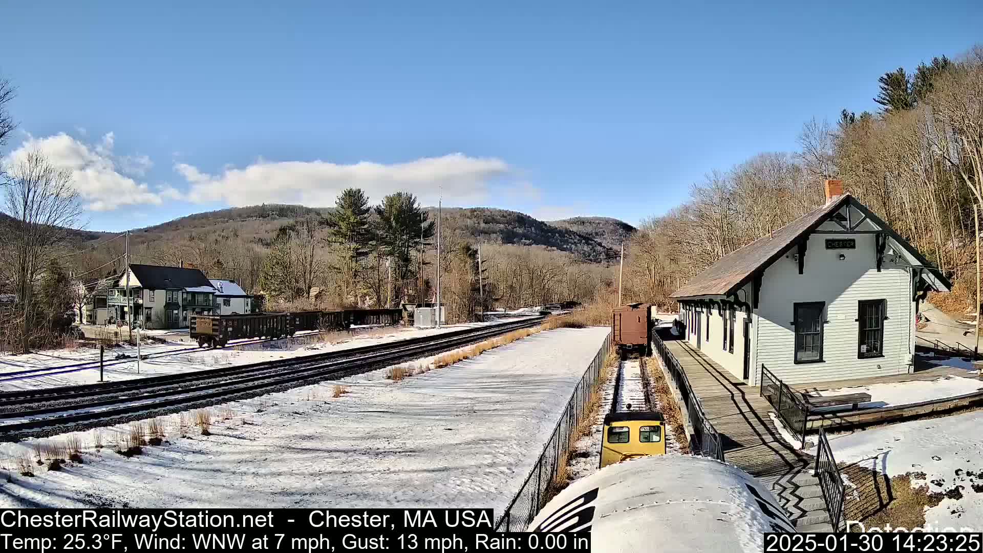 A snow-covered train station and tracks are seen on a partly sunny day with a background of hills and trees.