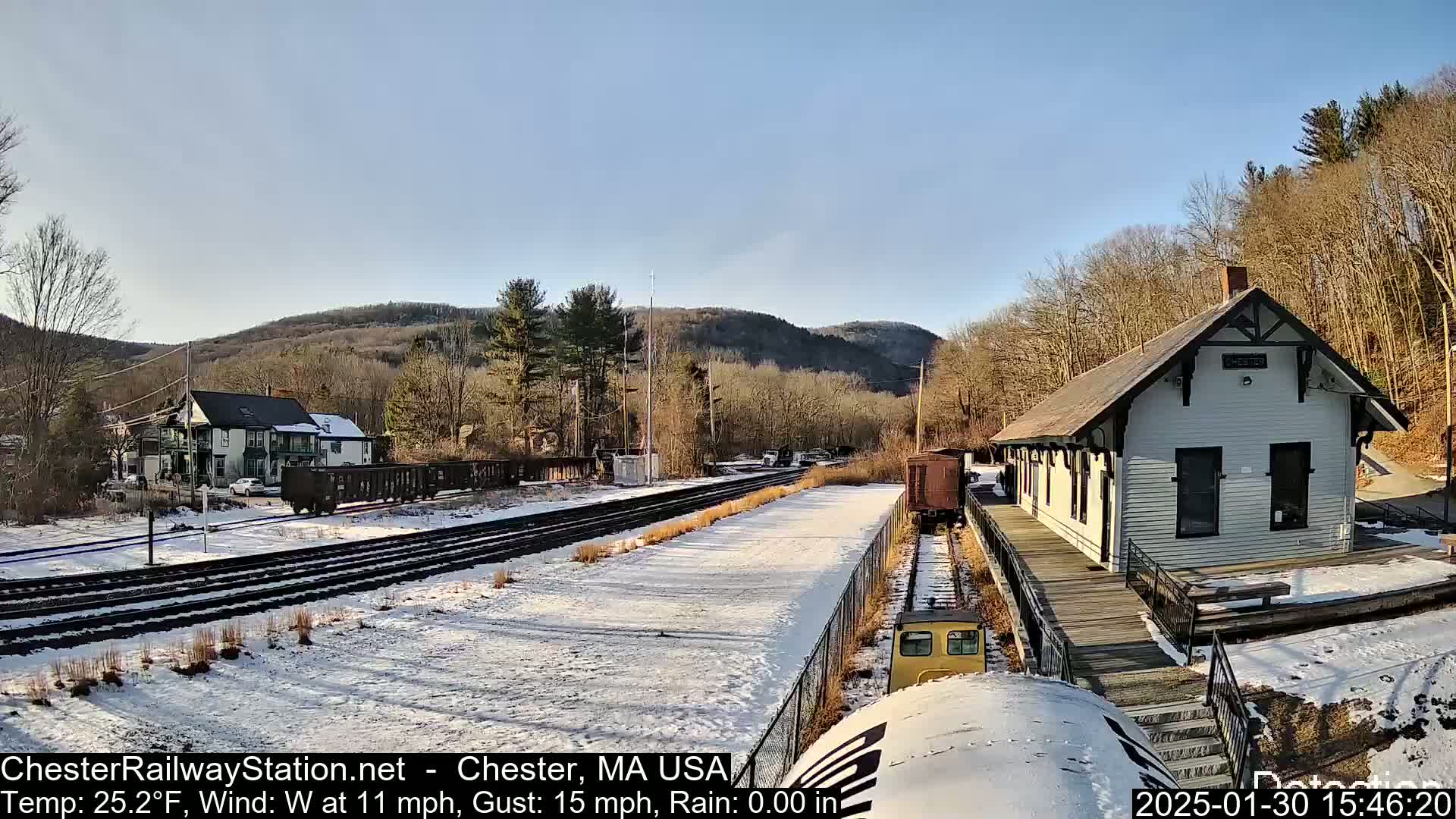 A snow-covered railway station and tracks sit beside a small white building, with a residential building and hills in the background under a clear, sunny sky.