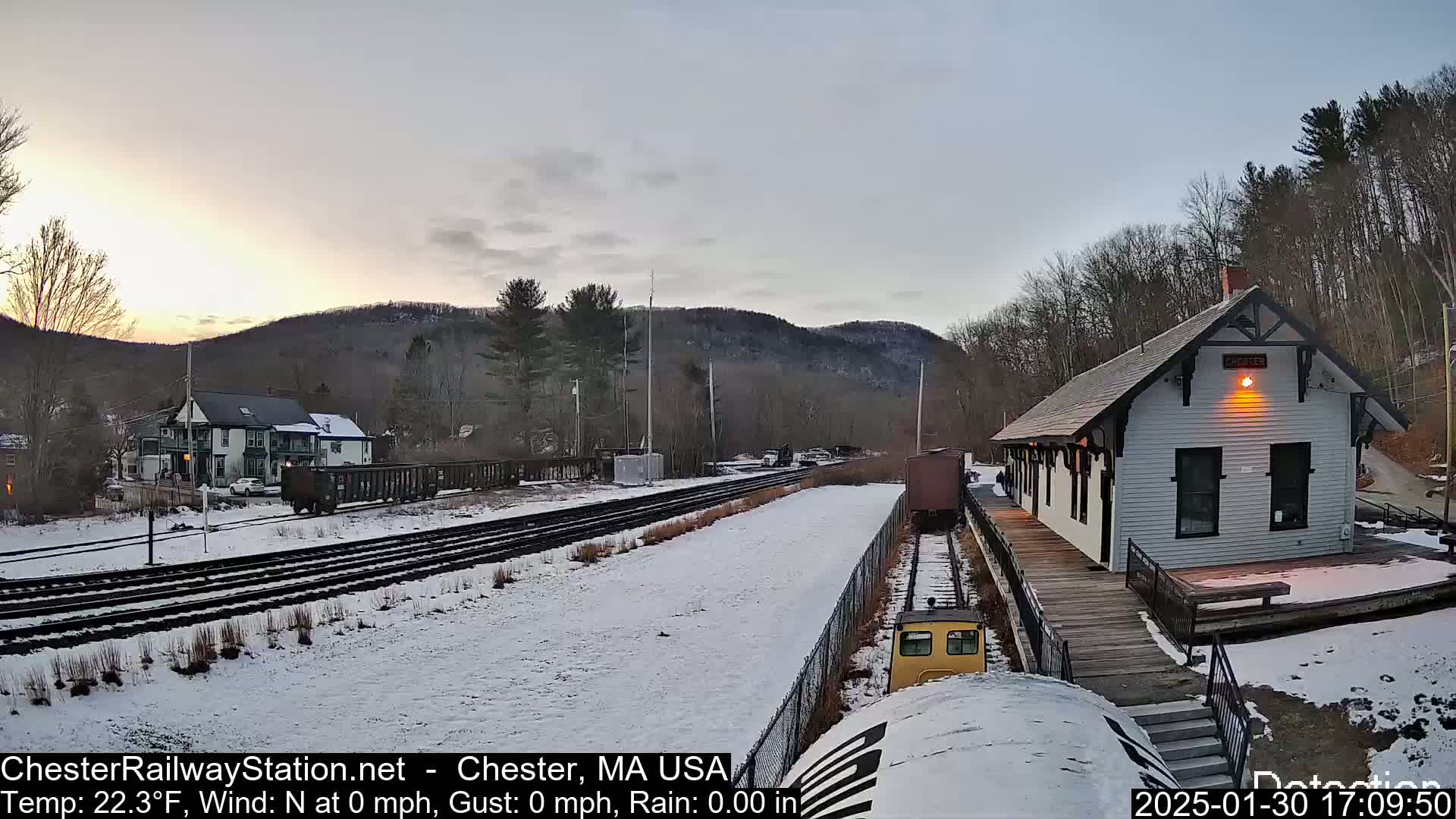 A snow-covered train station and tracks are situated at the base of a hill, under a twilight sky.