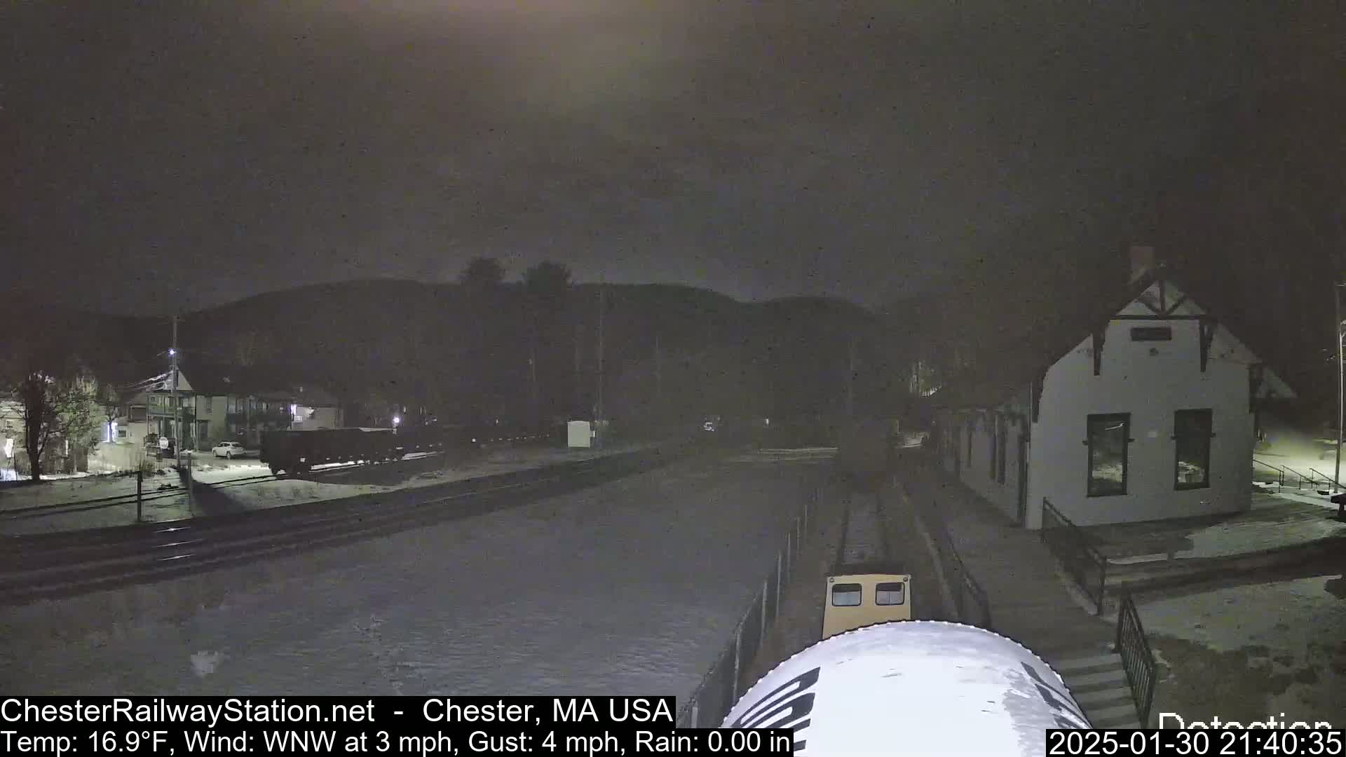 A nighttime view of a snow-covered train station and railway with a train approaching, under a dark sky with a temperature of 16.9°F and a wind blowing from the west-northwest at 3 mph with gusts up to 4 mph.