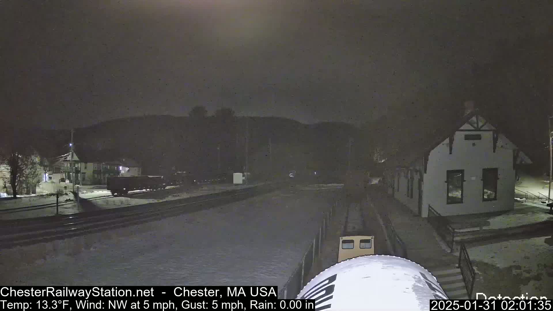 A nighttime view of a snow-covered train station and tracks with a train car partially visible, under a dark sky.