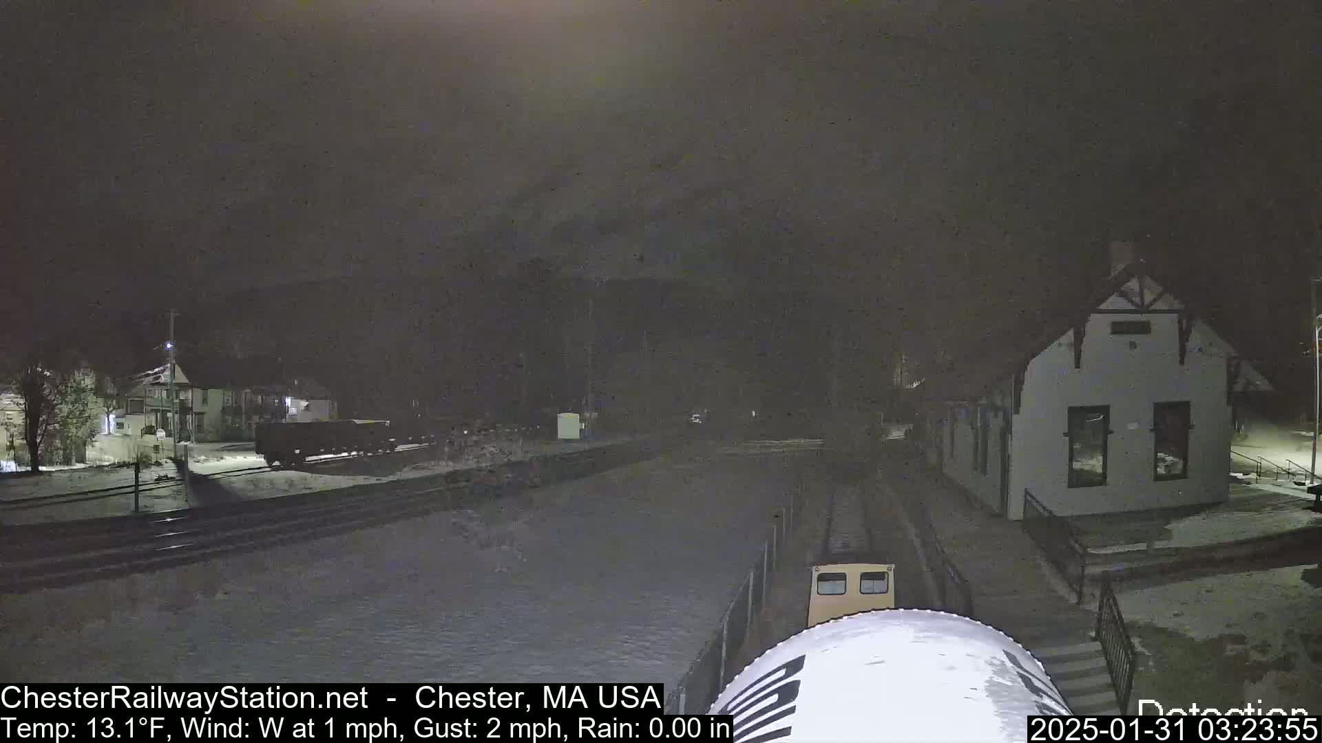 A nighttime view of a snow-covered train station and tracks, with a train partially visible in the foreground under a dark sky.
