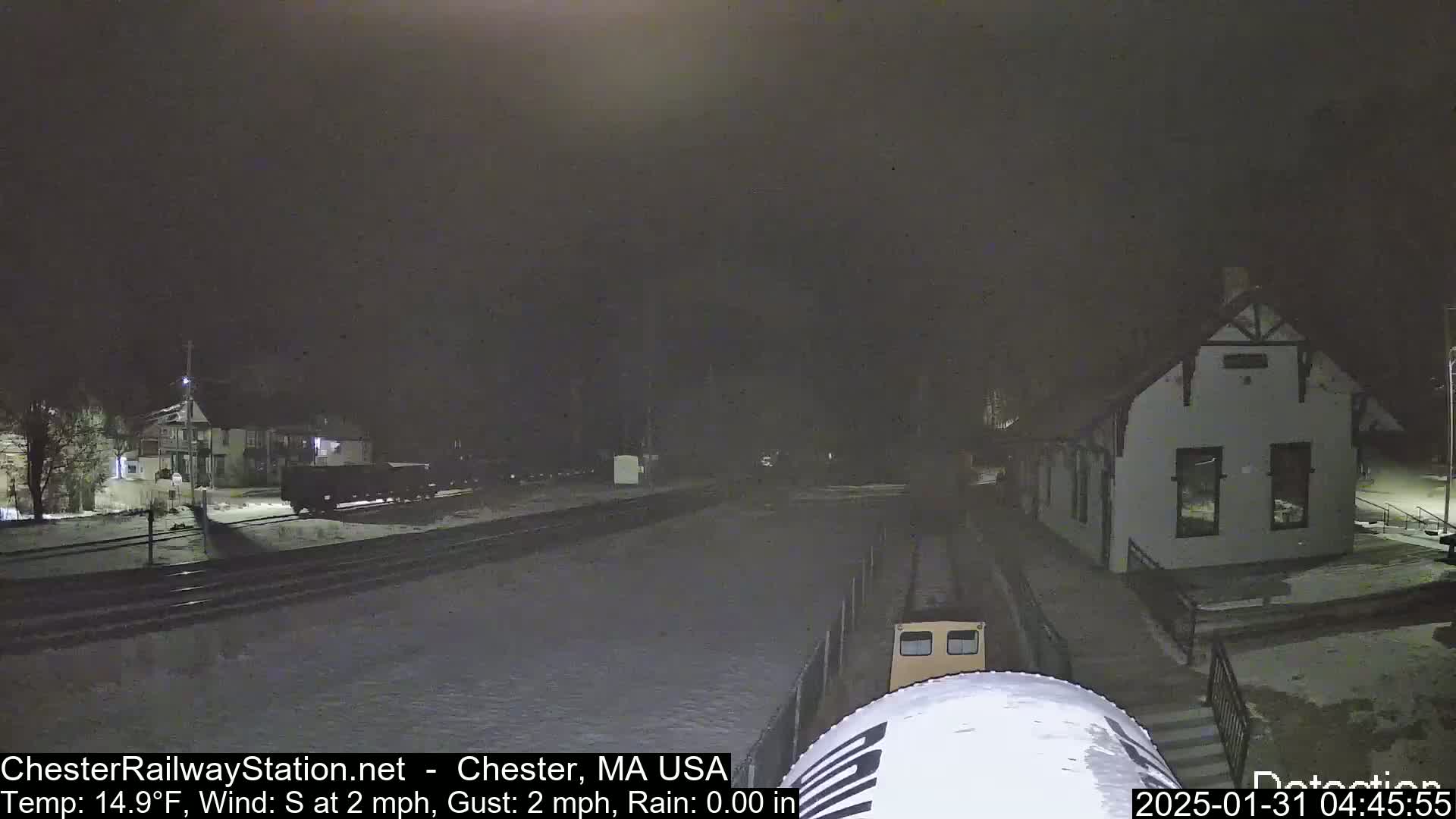 A nighttime view of a snow-covered train station and railway tracks, with a small train visible in the foreground, under a dark sky with a south wind.