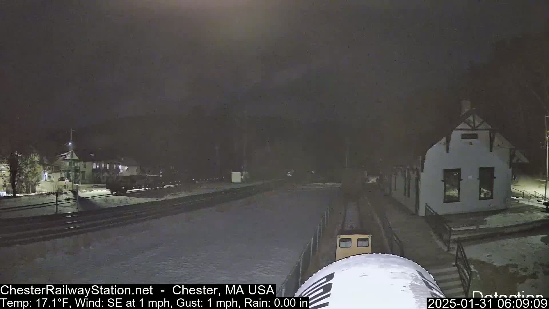 A nighttime view of a train approaching a snow-covered station building under a dark sky with a southeast wind of 1 mph and a temperature of 17 degrees Fahrenheit.