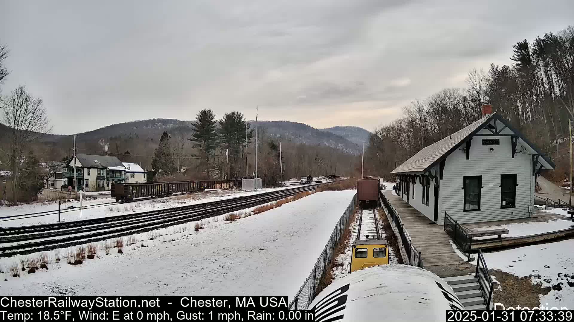 A snow-covered train station and railway tracks are seen in a winter landscape with overcast skies.