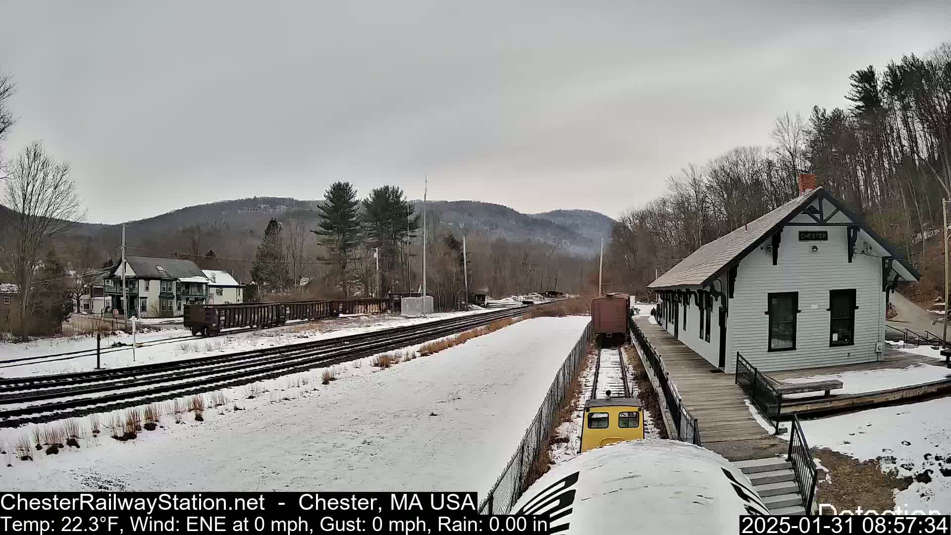 A snow-covered train station and tracks are visible, with a small yellow train car nearby, set against a backdrop of a snowy landscape and distant, wooded hills under an overcast sky.