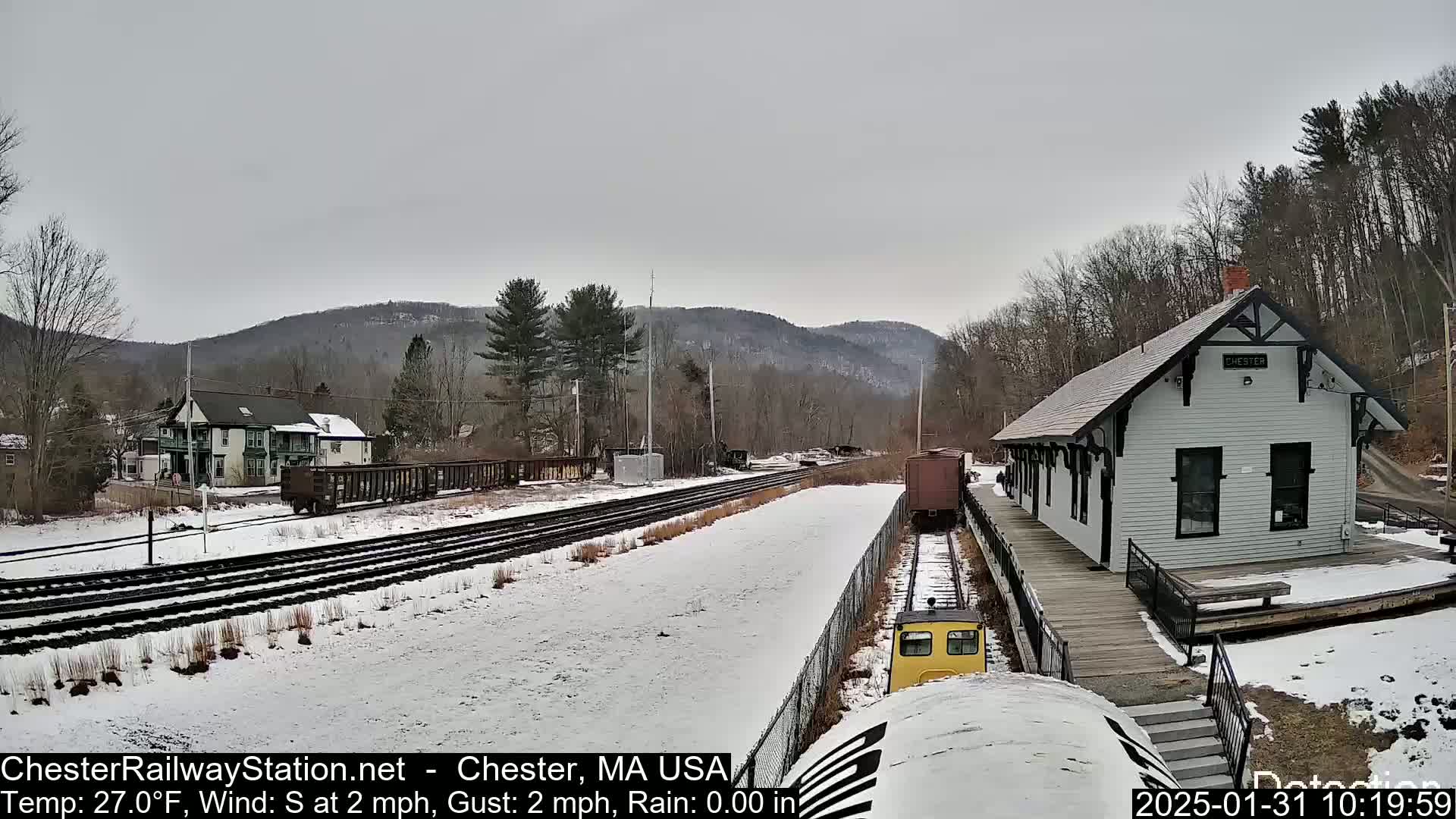 A snow-covered train station and tracks are visible, with a small yellow train car and freight cars in view under an overcast sky.