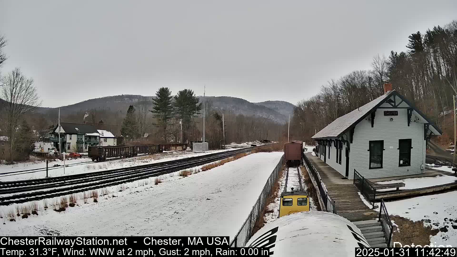 A snow-covered train station and railway tracks are visible, with a small yellow train car and freight cars nearby, under a gray, overcast sky.