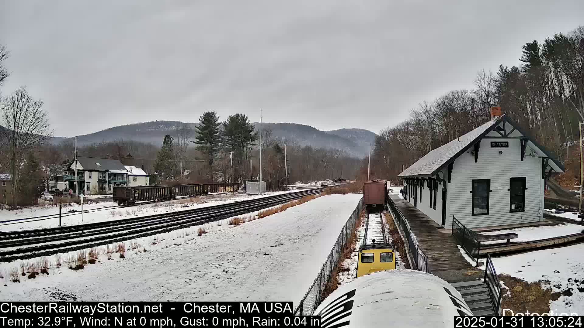 A snow-covered train station and tracks are visible under a cloudy sky, with a small yellow train car and freight cars present; light rain is falling.