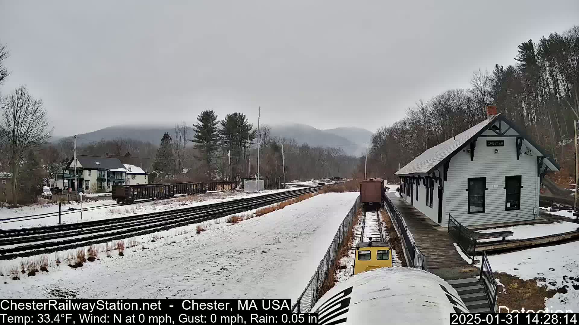 A snow-covered train station sits beside a train track in a hilly, wooded area under an overcast sky.