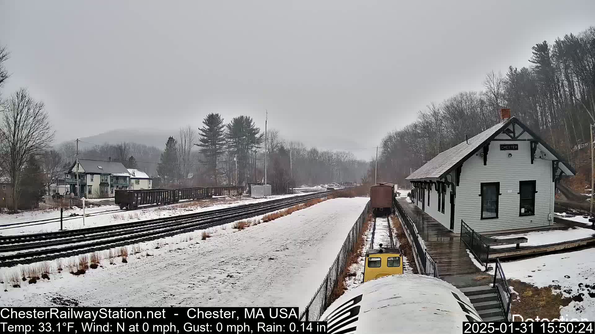 A snow-covered train yard with a small white train station and a freight train is visible under an overcast sky, with light rain.