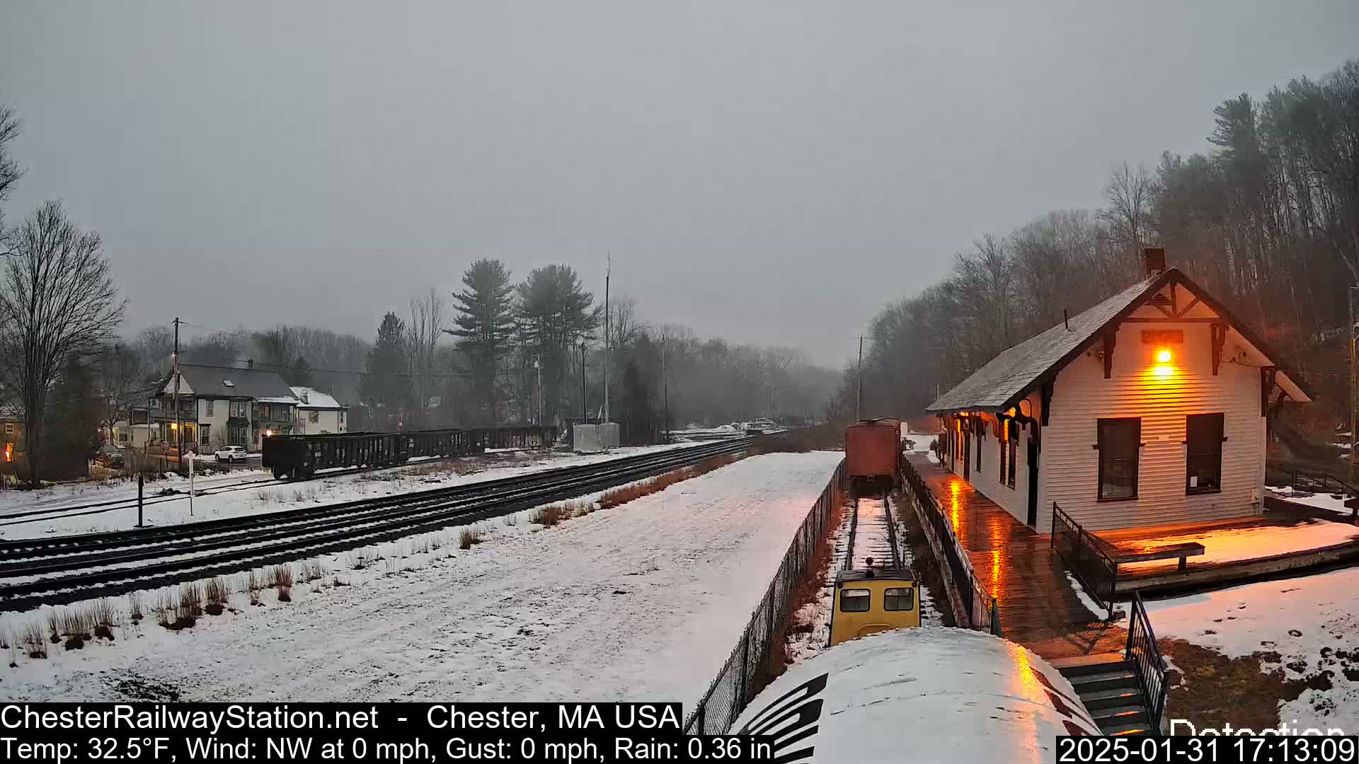 A snow-covered train station and tracks are visible under an overcast sky with light rain.