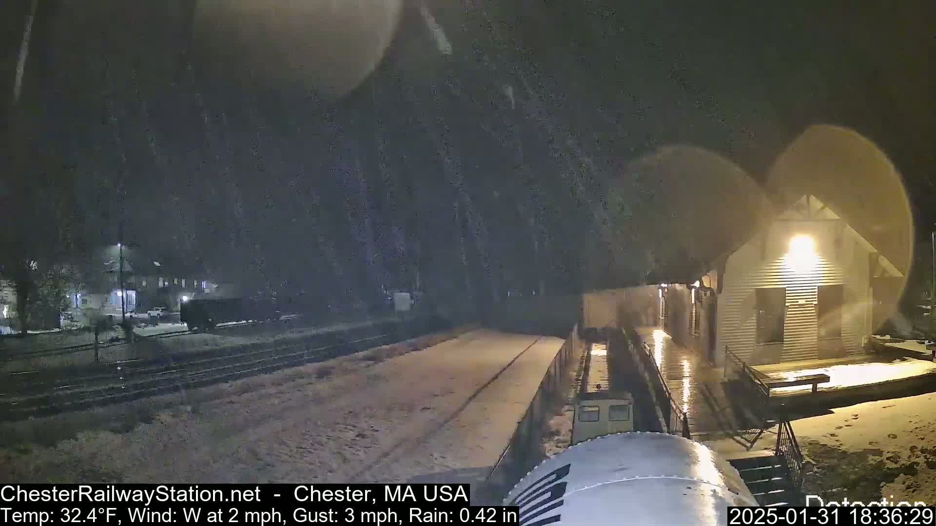 A snow-covered train platform and a small building are visible at night under falling snow and rain.