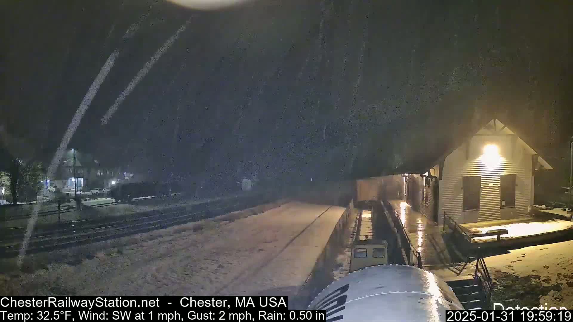 A snow-covered train platform and a small building are visible at night during a light snowfall with rain.
