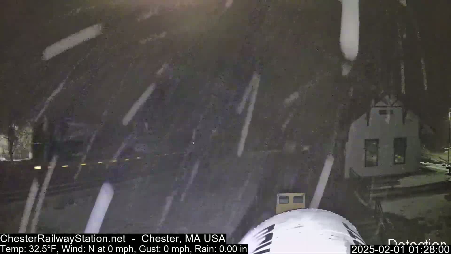 Snow falls heavily at night on a small building and a road.