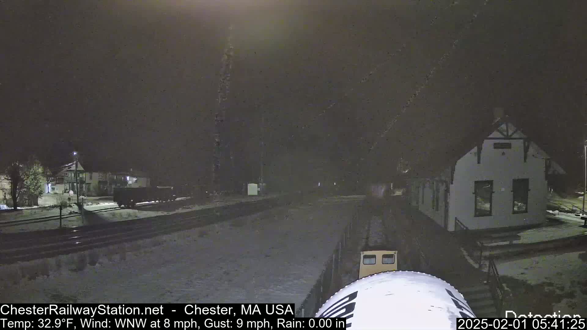 A nighttime, snowy scene shows a train passing a building near a railway track, under a dark sky with light rain.