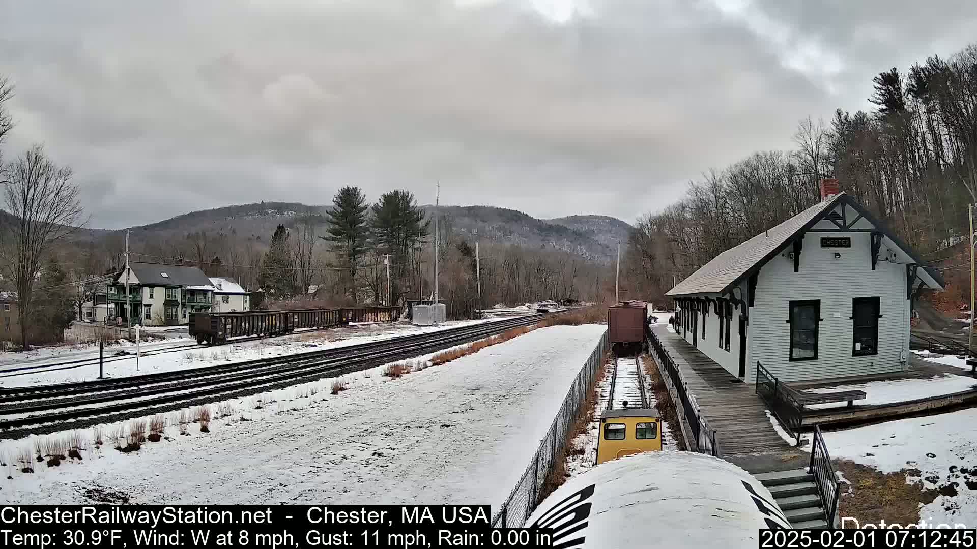 A snow-covered train station and tracks sit before a hilly landscape under an overcast sky.