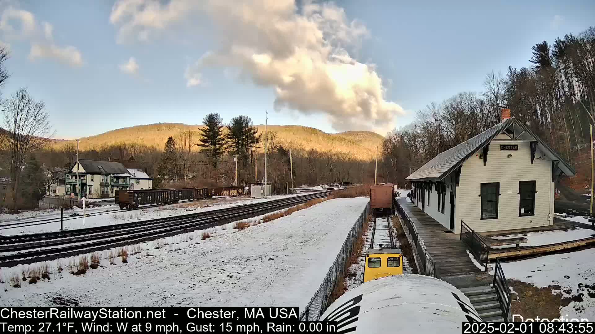 A snow-covered train yard with a small white train station, railway tracks, and freight cars, nestled amongst hills under a partly cloudy sky.
