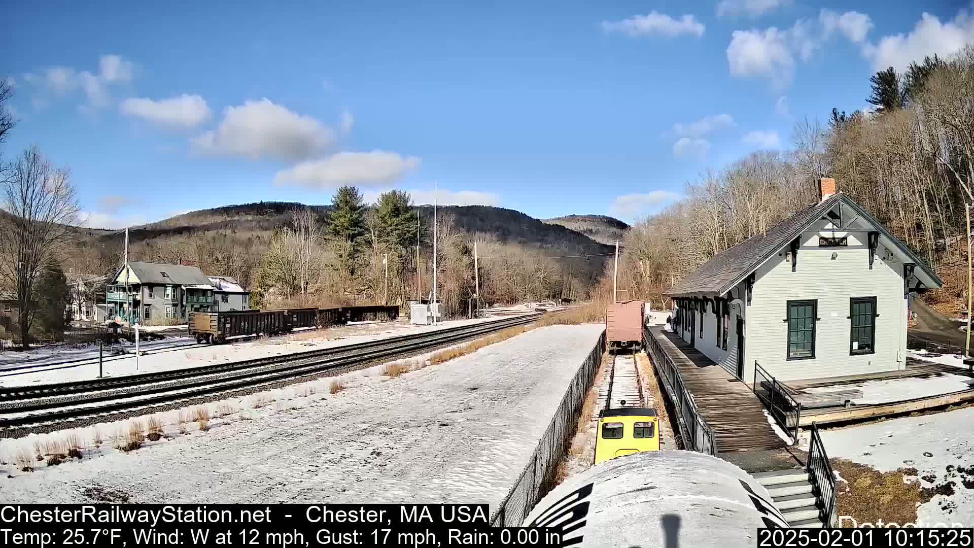 A train approaches a snow-covered train station on a sunny day with a partly cloudy sky and a westerly wind.