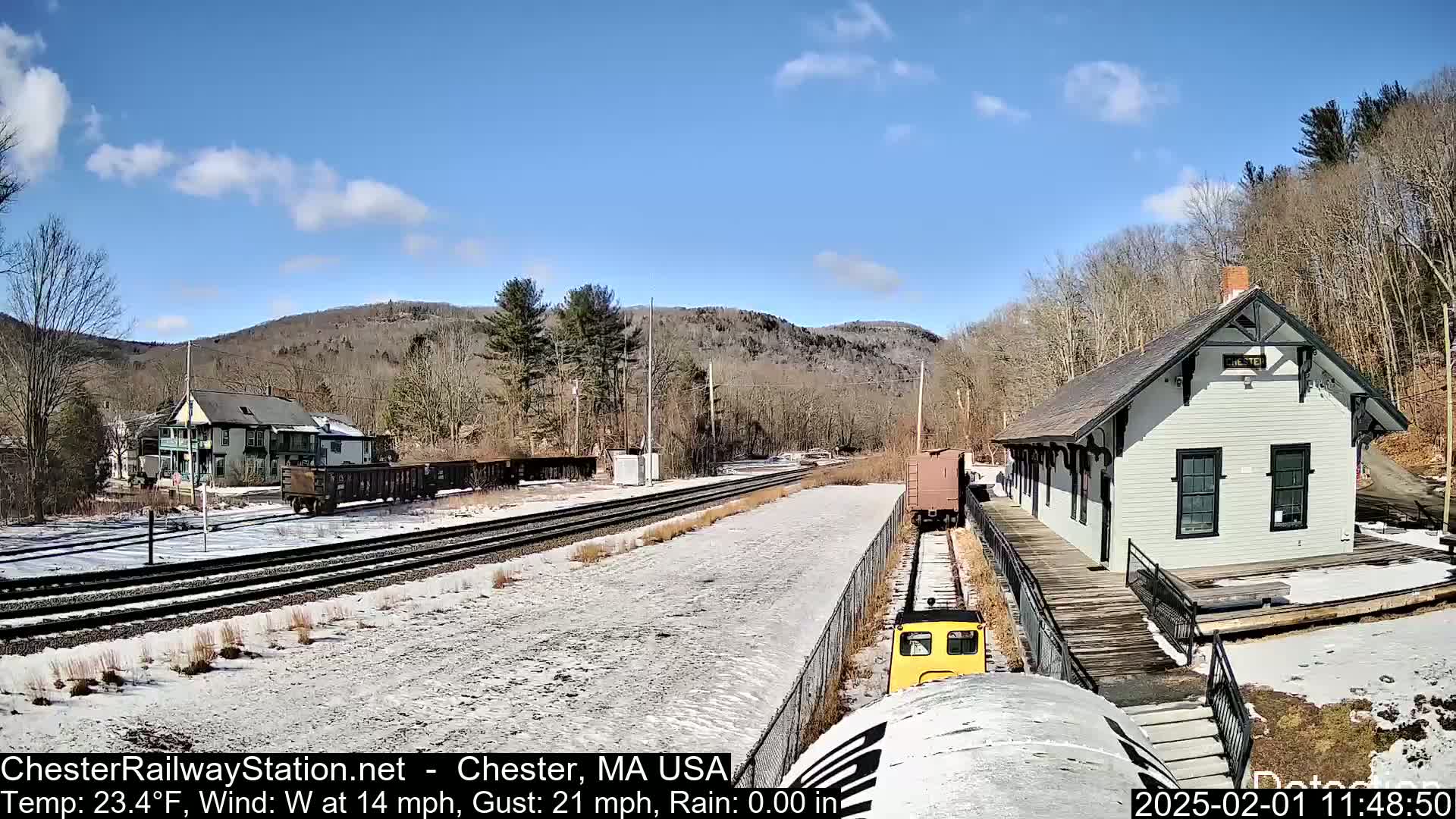 A small yellow train approaches a light-grey train station on a partly sunny day with snow on the ground, a light dusting of snow on the surrounding trees and buildings, and a backdrop of wooded hills.