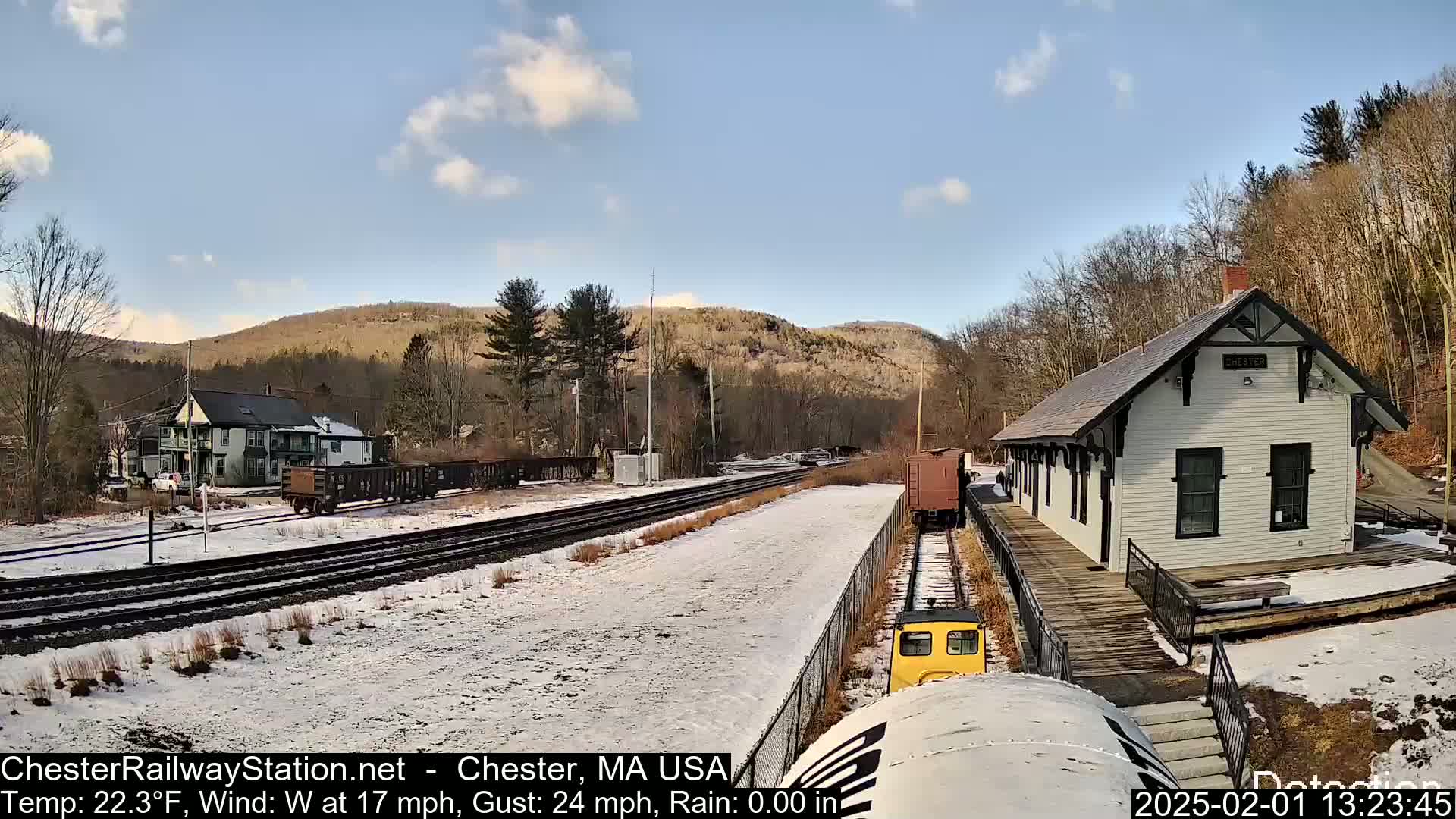 A snow-covered train station and tracks are visible, with a small train car nearby, under a partly cloudy sky, and hills in the background.