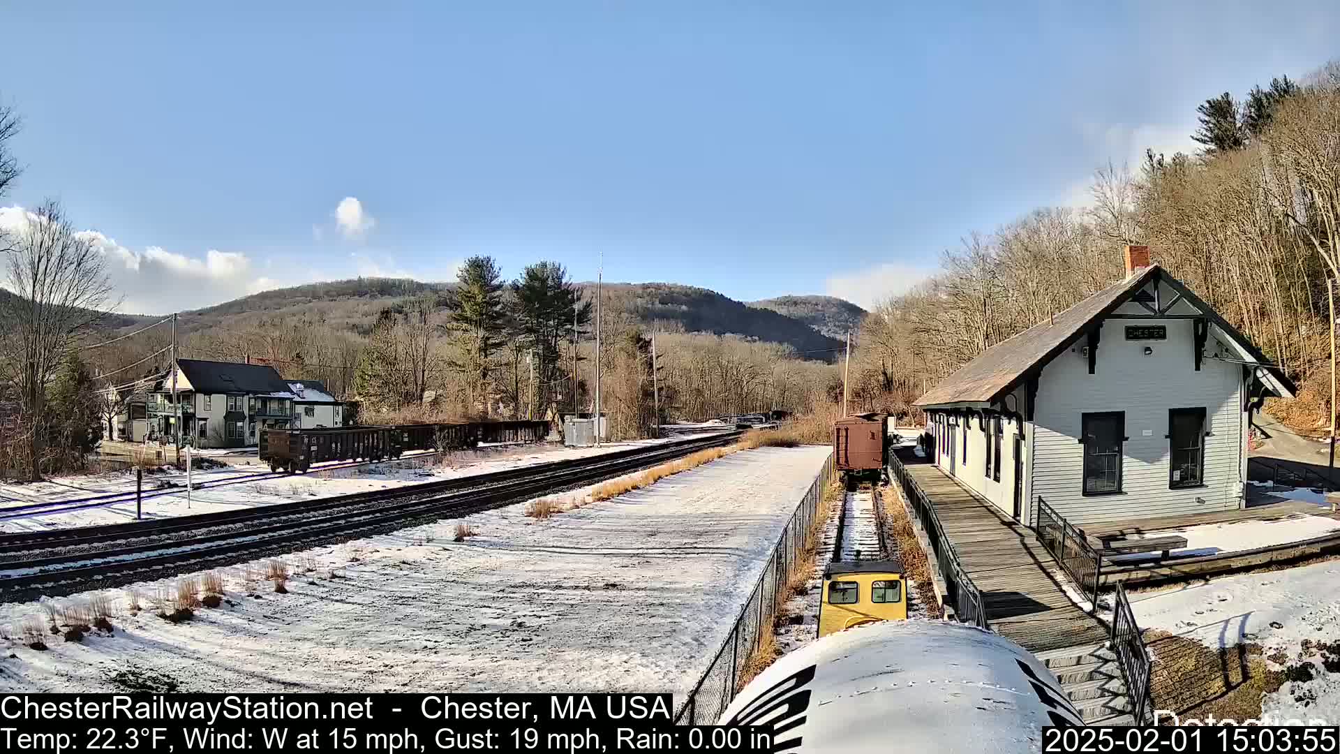 A train approaches a snow-covered railway station next to a two-story building, with a partly sunny sky and mountains in the background.
