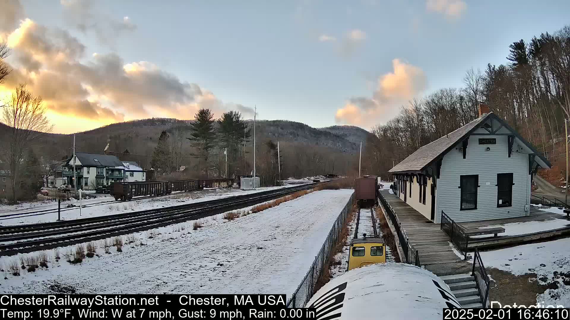 A snow-covered train station and railway tracks are seen at sunset under a partly cloudy sky, with hills in the background.